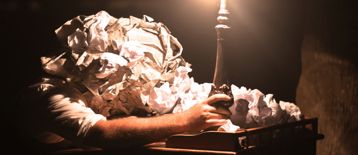 Crumpled sheets of paper piled around a typewriter beneath a desk lamp
