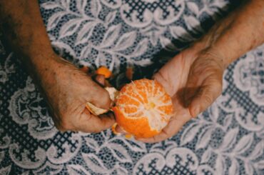 A close-up of elderly hands peeling a fresh orange on a patterned fabric background.