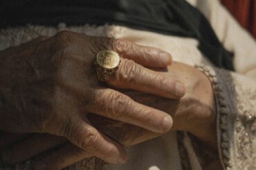 Close-up of an older person's hands, one wearing a gold ring, resting on embroidered cloth